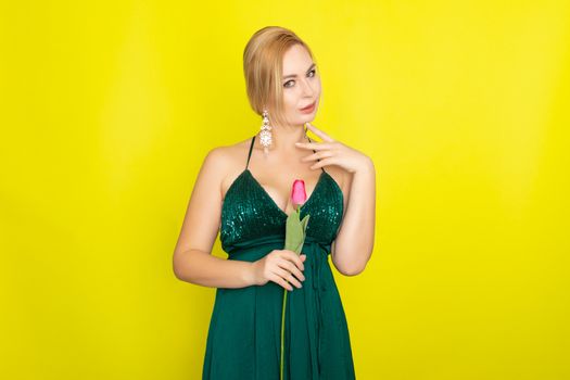 Blonde woman in green evening dress holding one tulip in her hands over yellow background