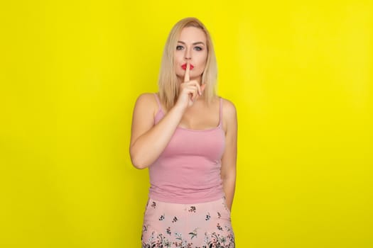 Indoor summer closeup portrait of young stylish fashion glamorous blonde woman posing in pink shorts and shirt, standing over yellow background