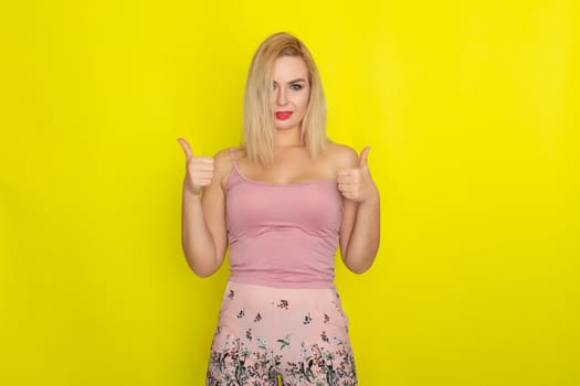 Indoor summer closeup portrait of young stylish fashion glamorous blonde woman posing in pink shorts and shirt, standing over yellow background