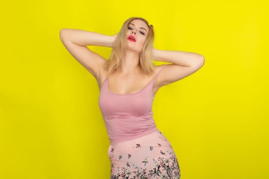 Indoor summer closeup portrait of young stylish fashion glamorous blonde woman posing in pink shorts and shirt, standing over yellow background