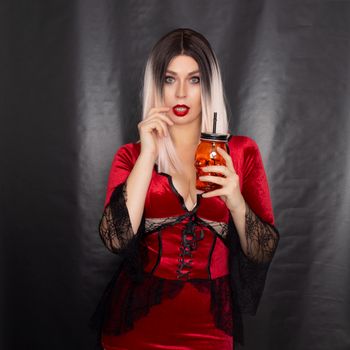 Young beautiful blonde woman in a red vampire dress holds an orange glass in the shape of a skull in her hands