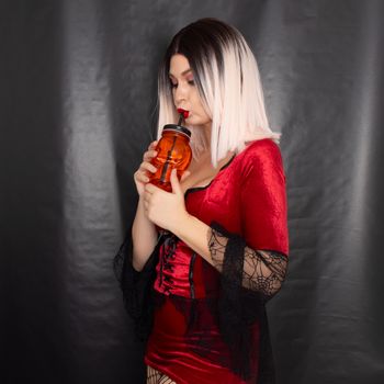 Young beautiful blonde woman in a red vampire dress holds an orange glass in the shape of a skull in her hands