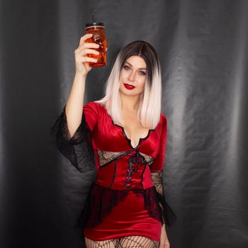 Young beautiful blonde woman in a red vampire dress holds an orange glass in the shape of a skull in her hands