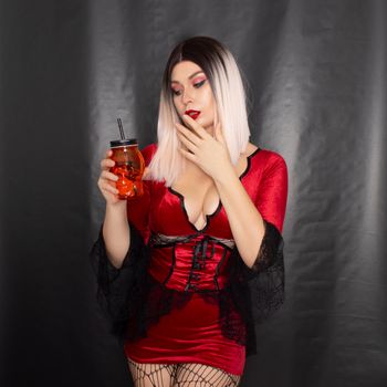 Young beautiful blonde woman in a red vampire dress holds an orange glass in the shape of a skull in her hands