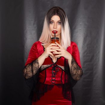 Young beautiful blonde woman in a red vampire dress holds an orange glass in the shape of a skull in her hands