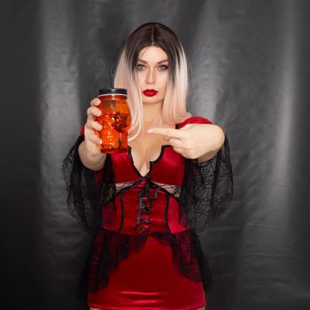 Young beautiful blonde woman in a red vampire dress holds an orange glass in the shape of a skull in her hands