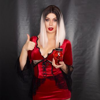 Young beautiful blonde woman in a red vampire dress holds an orange glass in the shape of a skull in her hands