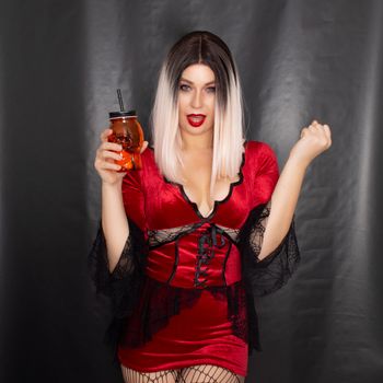 Young beautiful blonde woman in a red vampire dress holds an orange glass in the shape of a skull in her hands