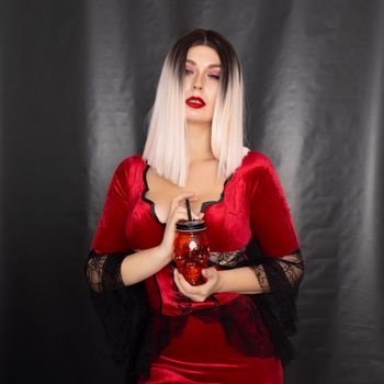 Young beautiful blonde woman in a red vampire dress holds an orange glass in the shape of a skull in her hands