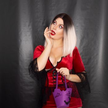 Young beautiful blonde woman in a red vampire dress holds a Halloween purple bag with bats for sweets