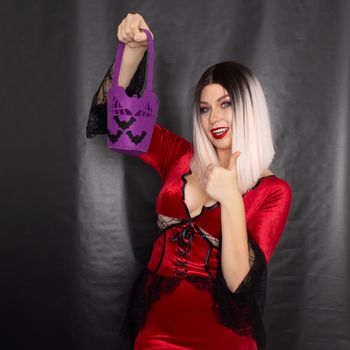 Young beautiful blonde woman in a red vampire dress holds a Halloween purple bag with bats for sweets