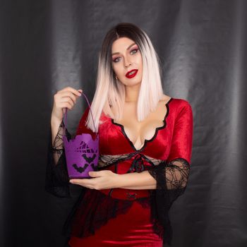 Young beautiful blonde woman in a red vampire dress holds a Halloween purple bag with bats for sweets