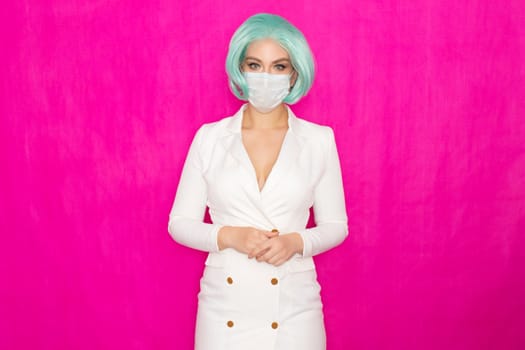 Beautiful young woman with short blue hair in a white business dress jacket with a medical mask posing on a pink background in the studio