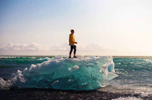 Tourist standing on an iceberg at the Diamond Beach in Jokulsarlon Glacier Lagoon in Iceland. This beach is known for its large ice rocks lying on the lava black sand.