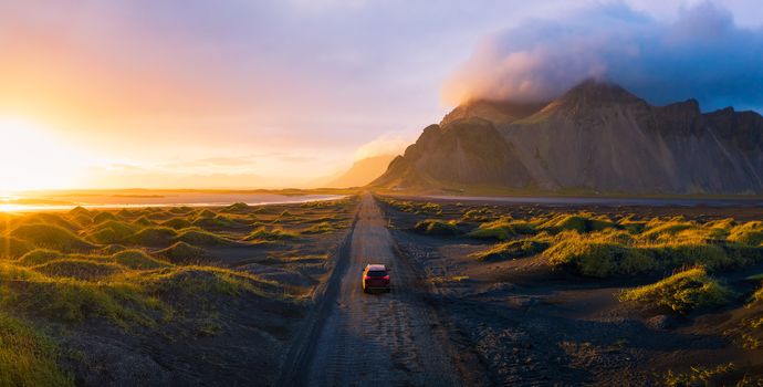 Panorama of a gravel road at a golden Sunset with Vestrahorn mountain in the background and a car driving the road in Iceland