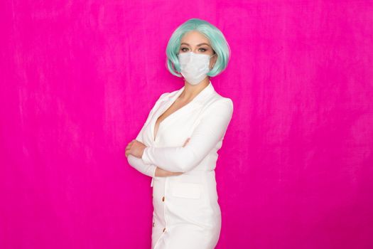 Beautiful young woman with short blue hair in a white business dress jacket with a medical mask posing on a pink background in the studio