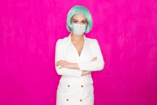 Beautiful young woman with short blue hair in a white business dress jacket with a medical mask posing on a pink background in the studio