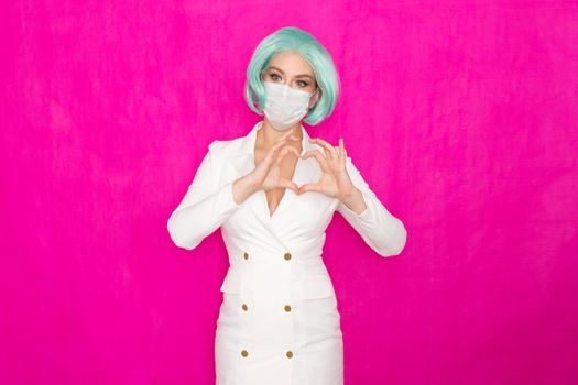 Beautiful young woman with short blue hair in a white business dress jacket with a medical mask posing on a pink background in the studio