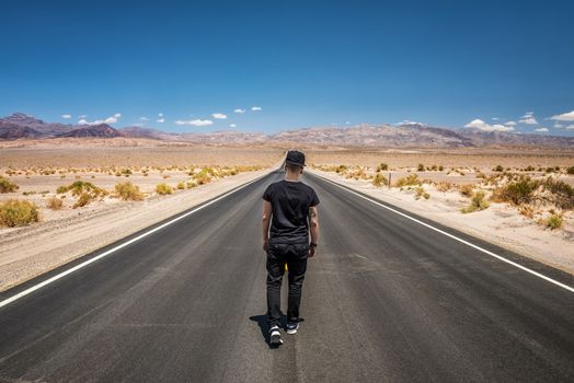 Young man walking alone through an empty street in the desert of Death Valley National Park on a bright sunny day. Loneliness, sadness , depression and solitude concept in nature.