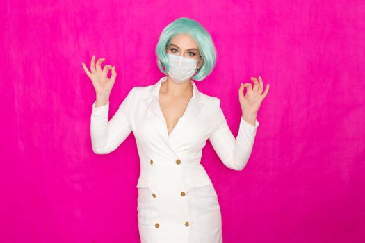 Beautiful young woman with short blue hair in a white business dress jacket with a medical mask posing on a pink background in the studio