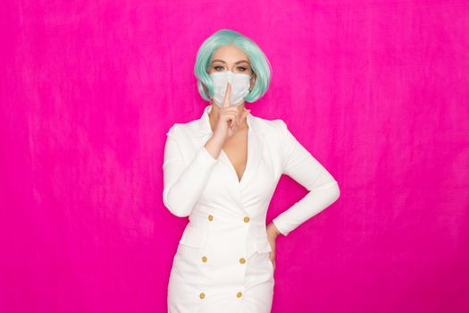 Beautiful young woman with short blue hair in a white business dress jacket with a medical mask posing on a pink background in the studio