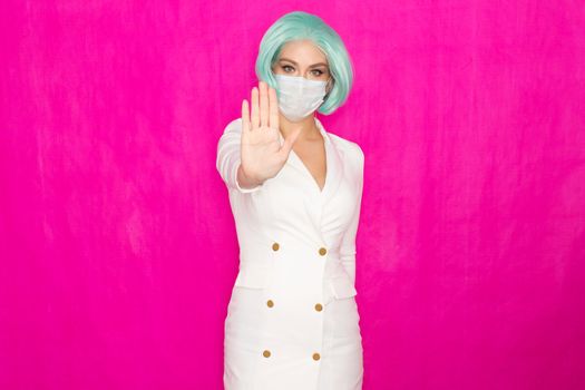 Beautiful young woman with short blue hair in a white business dress jacket with a medical mask posing on a pink background in the studio