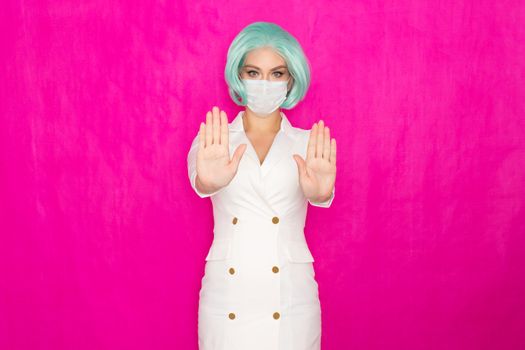 Beautiful young woman with short blue hair in a white business dress jacket with a medical mask posing on a pink background in the studio