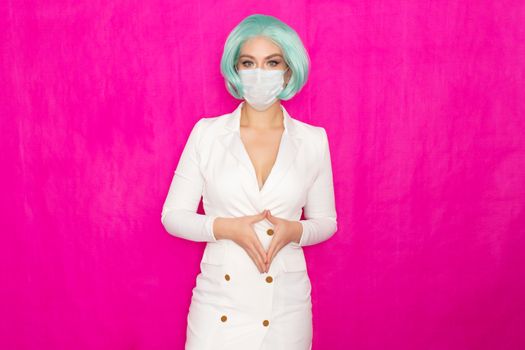 Beautiful young woman with short blue hair in a white business dress jacket with a medical mask posing on a pink background in the studio