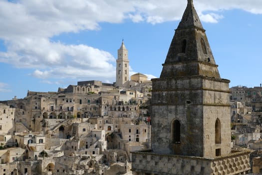 Point of two bell towers and church in the ancient city of Matera in Italy. Construction with blocks of tufa stone. Chileo blue with clouds.