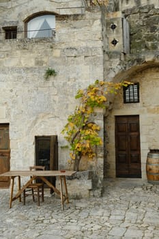 Matera, Basilicata, Italy. About 11/2019. Vine plant grown in a stone planter in the Sassi of Matera. Courtyard of a house with a wooden table and a wine barrel.
