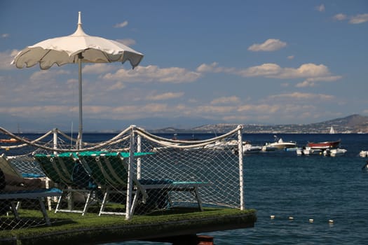 Ischia, Naples, Italy, About the July 2019.  Umbrella on a pier with a white parapet on the sea of Ischia. In the background a ferry boat.