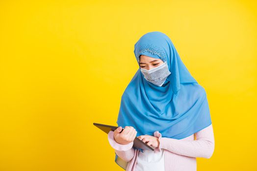 Asian Muslim Arab, Portrait of happy beautiful young woman Islam religious wear veil hijab and face mask protect she quarantines disease coronavirus touch on tablet screen isolated yellow background