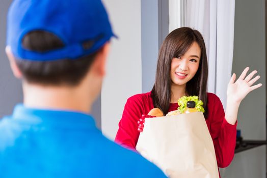 Asian young delivery man in blue uniform making grocery service giving fresh vegetables and fruits and food in paper bag to woman customer at front house after pandemic coronavirus, Back to new normal