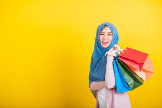 Asian Muslim Arab, Portrait of happy beautiful young woman Islam religious wear veil hijab funny smile she holding colorful shopping bags, studio shot isolated on yellow background