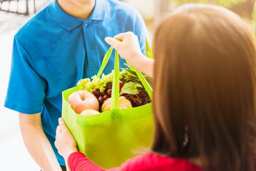 Asian young delivery man in uniform making grocery service giving fresh vegetables and fruits and food in green cloth bag to woman customer at door house after pandemic coronavirus, Back to new normal