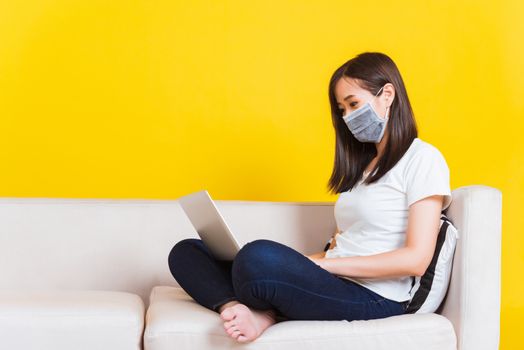 Portrait Asian of beautiful young woman sitting on sofa wearing medical face mask protective she work from home with laptop computer during Coronavirus studio shot isolated on yellow background