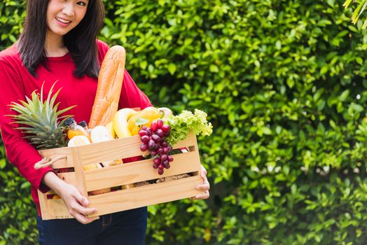 Portrait of Asian beautiful young woman farmer standing she smile and holding full fresh food raw vegetables fruit in a wood box in her hands on green leaves background