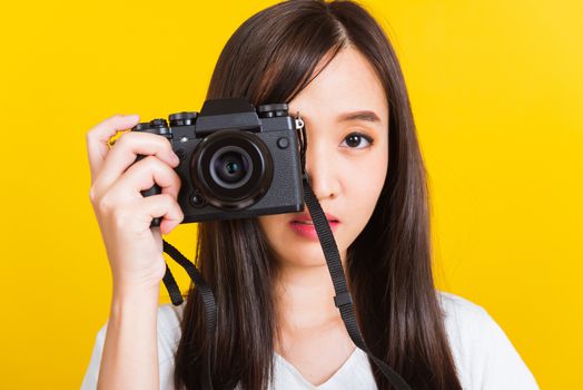 Portrait of happy Asian beautiful young woman photographer taking a picture and looking viewfinder on retro digital mirrorless photo camera ready to shoot, studio shot isolated on yellow background