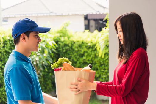 Asian young delivery man in uniform making grocery fast service giving fresh food in paper bag to woman customer receiving at house door under pandemic coronavirus, Back to new normal concept