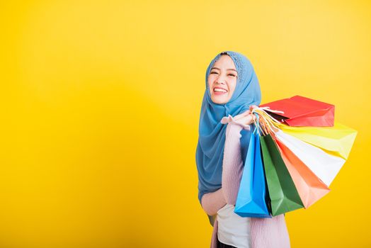 Asian Muslim Arab, Portrait of happy beautiful young woman Islam religious wear veil hijab funny smile she holding colorful shopping bags so glad shopping bags hand raise in studio isolated on yellow