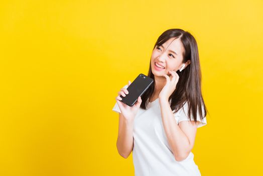 Portrait Asian of a happy beautiful young woman holding a mobile smart phone and wearing wireless headphones listening to music from smartphone studio shot isolated on yellow background