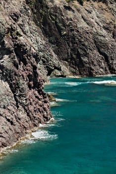 Sea waves break on the rocks of the Ligurian mountain.  Near the Cinque Terre a seascape with blue sea and dark red rocks. Village of Framura.