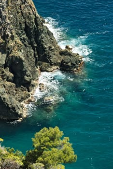 Sea waves break on the rocks of the Ligurian mountain.  Near the Cinque Terre a seascape with blue sea and dark red rocks. Village of Framura.