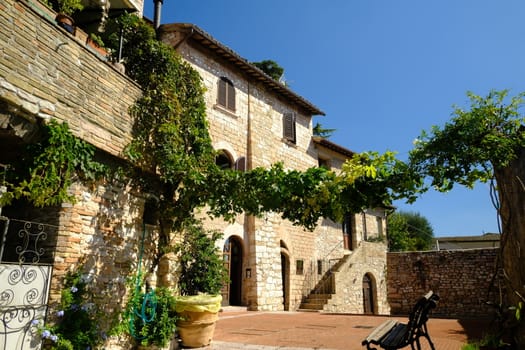 Alley of the city of Assisi with stone facades of historic houses. Arbor with climbing vine plant and flags on the walls of buildings.