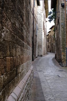 Alley of the city of Assisi with stone facades of historic houses. 	Narrow street of the city with the walls of the stone houses. Deserted road.