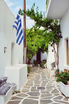 Picturesque narrow street with traditional whitewashed houses with greek flag of Naousa town in famous tourist attraction Paros island, Greece