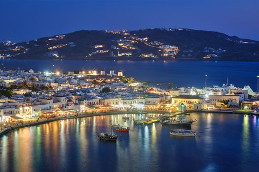 View of Mykonos Chora town Greek tourist holiday vacation destination with famous windmills, and port with boats and yachts illuminated in the evening blue hour . Mykonos, Cyclades islands, Greece
