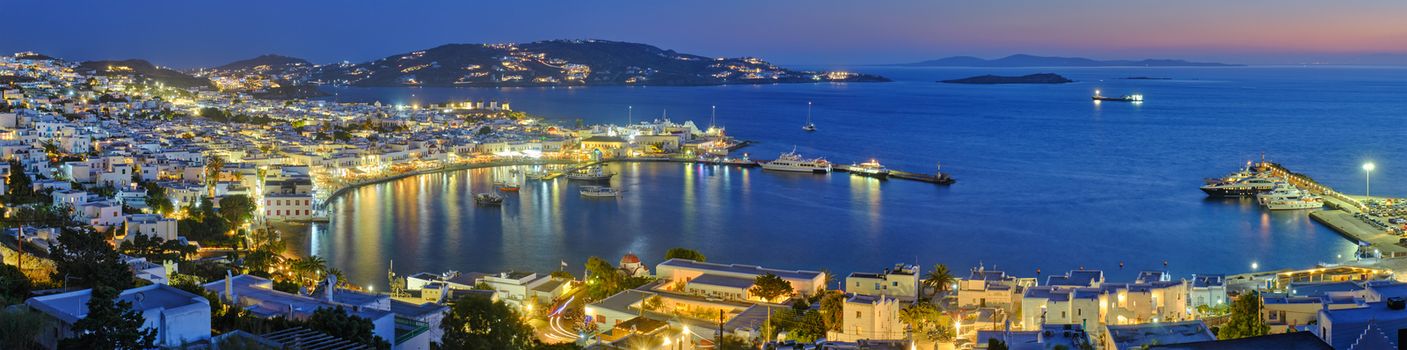 Panorama of Mykonos town Greek tourist holiday vacation destination with famous windmills, and port with boats and yachts illuminated in the evening blue hour . Mykonos, Cyclades islands, Greece