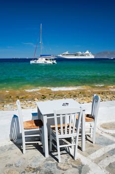 Tourist greek scene - restaurant cafe table on quay promenade with catamaran yacht and cruise liner and Aegean sea in background on beautiful summer day. Chora, Mykonos island, Greece