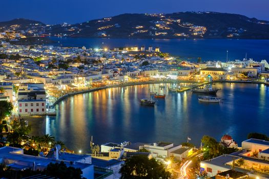 View of Mykonos town Greek tourist holiday vacation destination with famous windmills, and port with boats and yachts illuminated in the evening blue hour . Mykonos, Cyclades islands, Greece